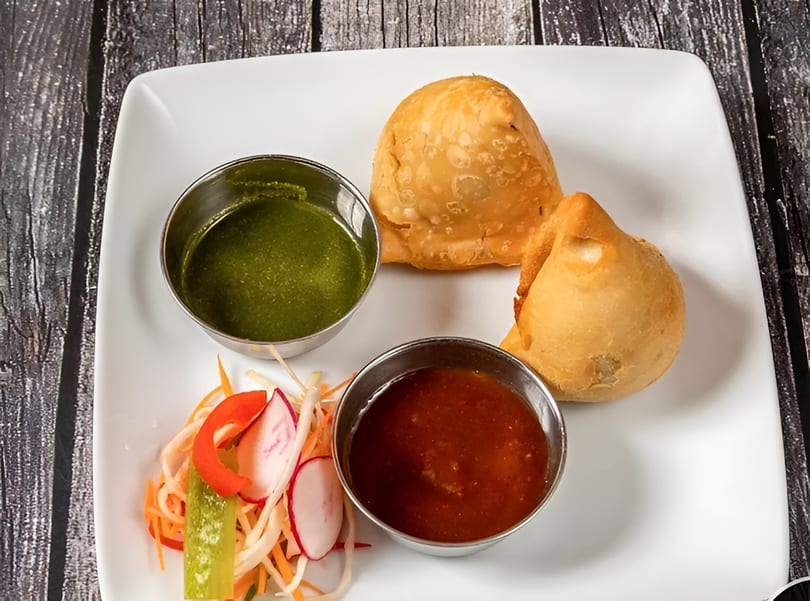 Two golden-brown samosas with green and red dipping sauces and a side salad on a white plate.