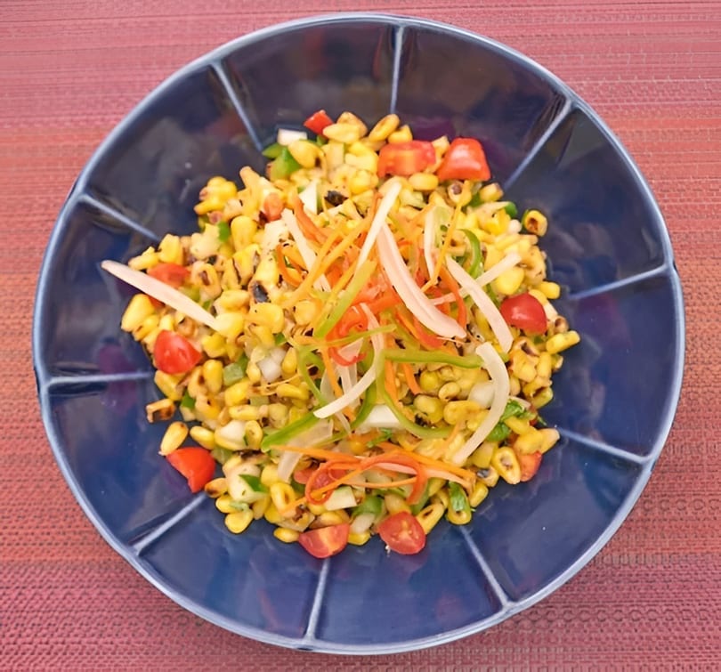 Grilled corn salad with cherry tomatoes, bell peppers, onions, and shredded carrots in a blue bowl.