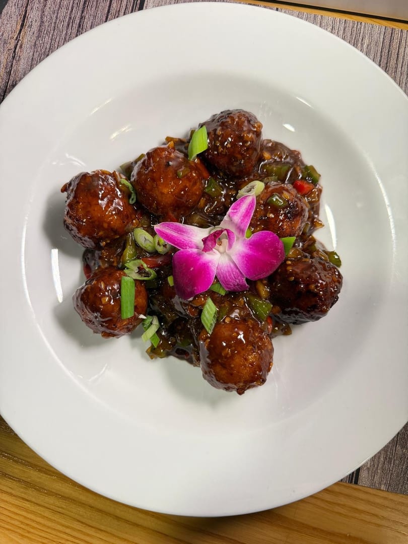 Brown meatballs in a dark sauce with green onions and a purple orchid on a white plate.