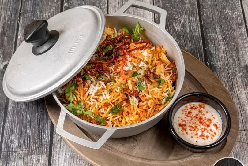 Biryani with fried onions and cilantro in a pot with a side of raita on a wooden tray.