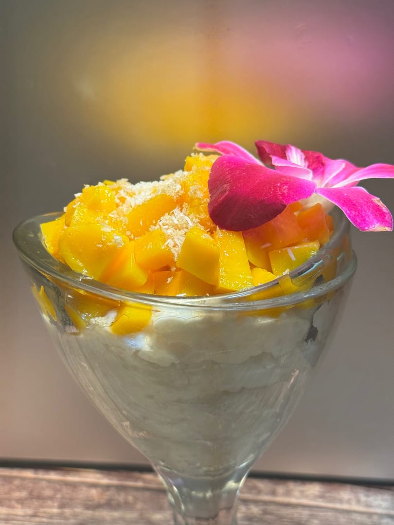 A glass of mango sticky rice dessert, topped with shredded coconut and a pink orchid.