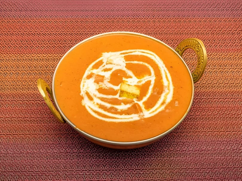 A golden bowl of creamy orange butter chicken, garnished with white cream swirls and paneer.