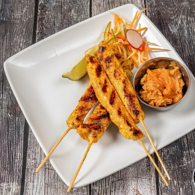 Grilled chicken satay skewers with peanut sauce, lime, and shredded vegetables on a white plate.